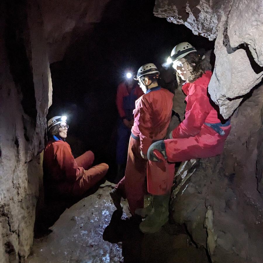 A group of people in red caving suits are gathered under the sloping ceiling of a cave. They’re looking at each other, lit only by their head torches.  