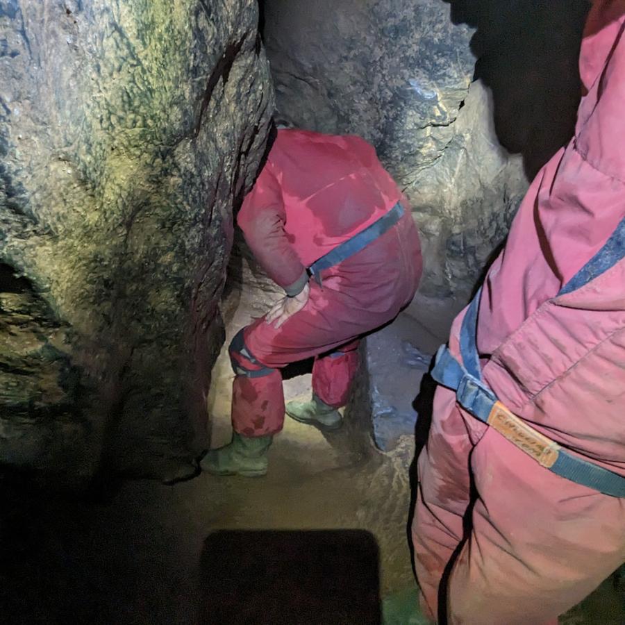 A person in a red caving suit sticks their head inside a gap in the rock. We can’t see their head, only the rest of their body is visible.  