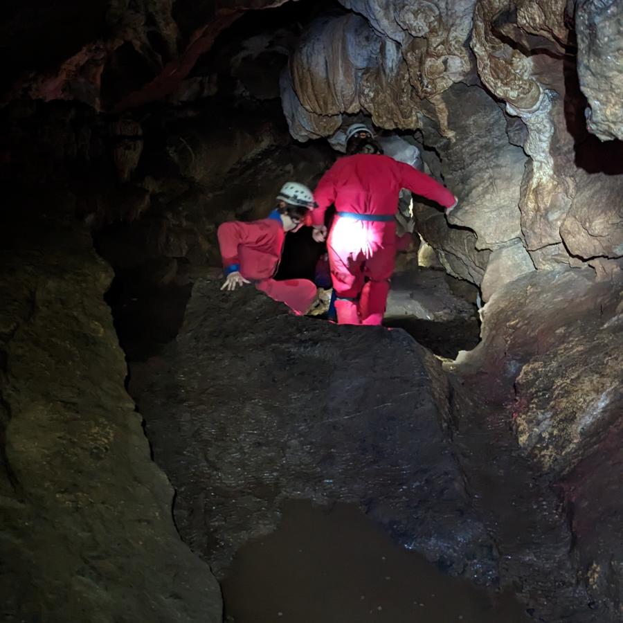 Two people in red caving suits making their way through a cave chamber. They’re at the very centre of the image, surrounded by rock. 