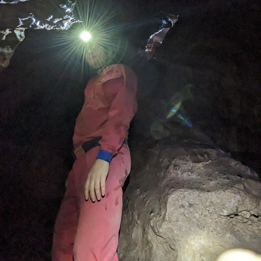 A person in a red caving suit looking directly at the camera in a cave. Their head torch is causing a lens flare.  