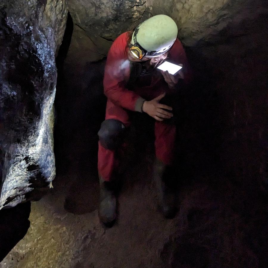 A person in a red caving suit leaning against a cave wall, listening to something on their phone. 