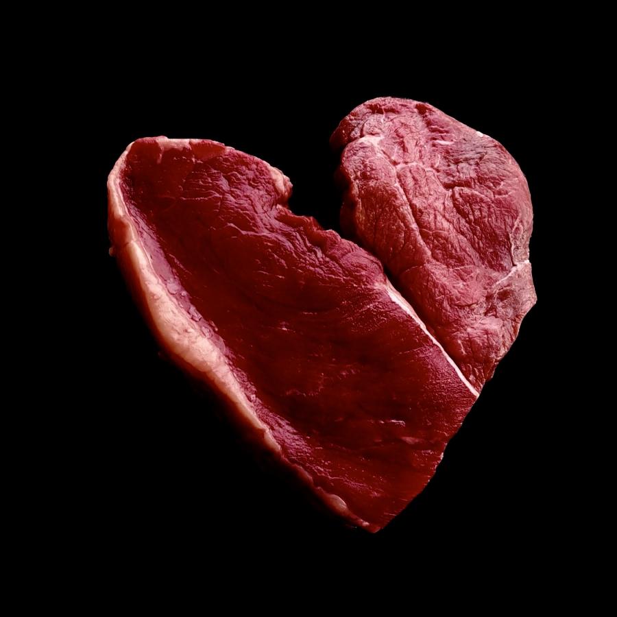 A piece of raw steak in the shape of heart sits in a black void. 