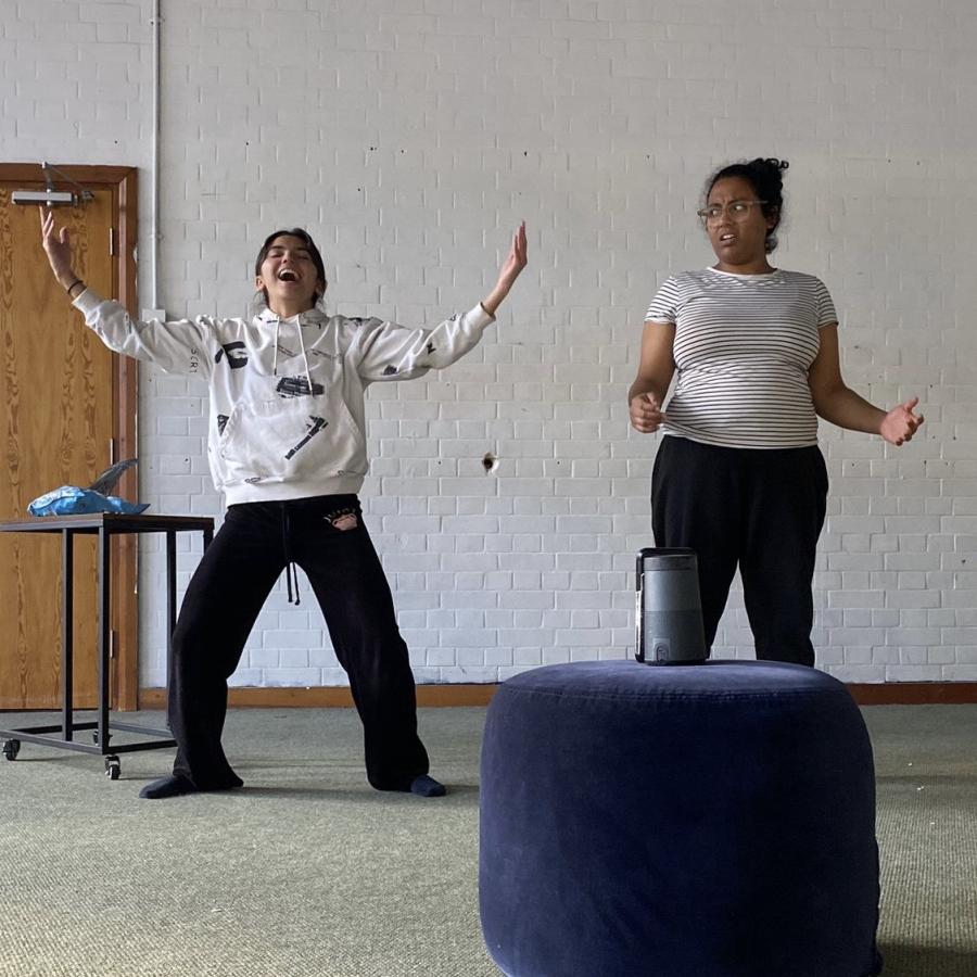 Two performers in the center of a white walled room. The performer on the left is laughing while throwing their arms open in a semicircle shape. The performer on the right looks at the left performer with a look of confusion. A blue ottoman sits in the co 