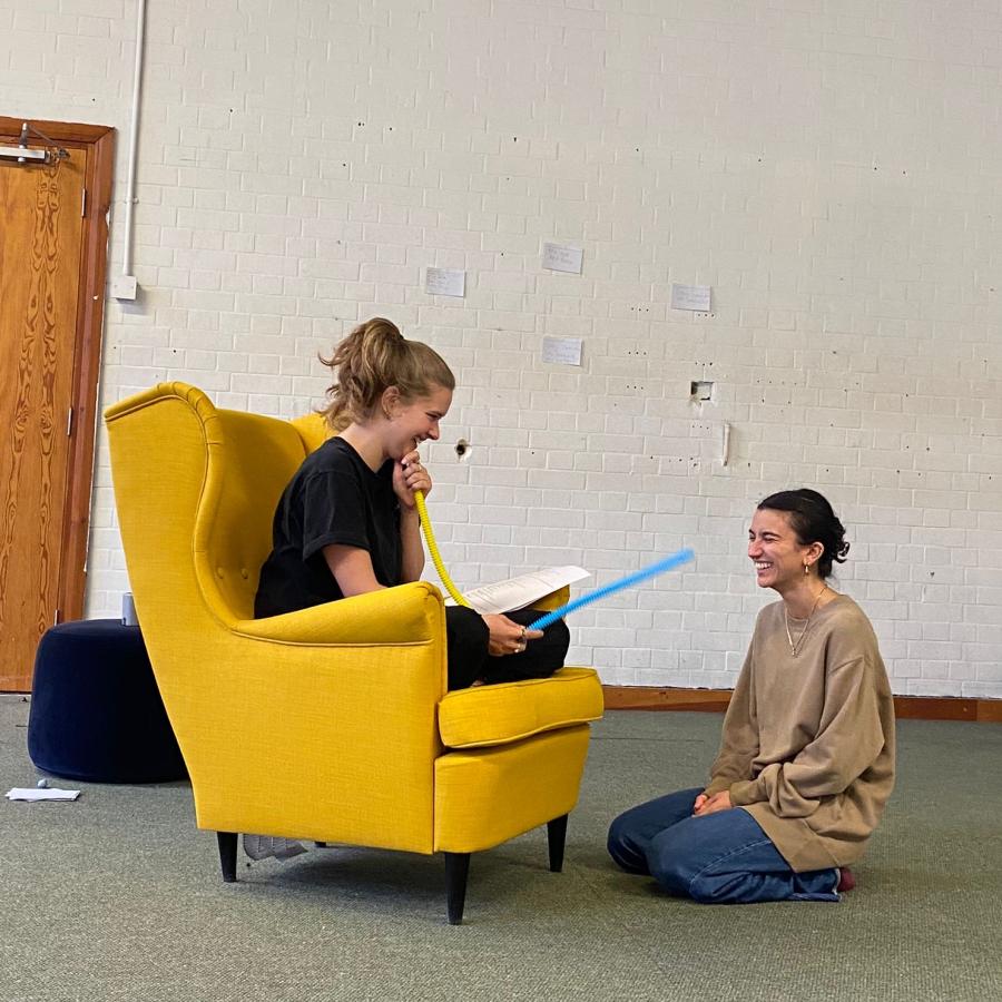 Two performers in a white walled room. The performer on the right sits cross legged on a yellow armchair looking to the performer on the right, who is sitting cross legged on the ground. Both performers are laughing. 