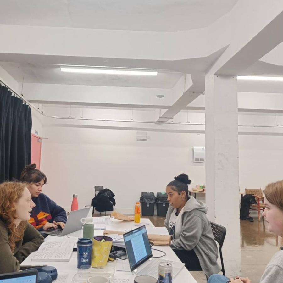 Four young women sitting at a table with papers, laptops, and waterbottles. They are in a white room with black curtains on the wall.  