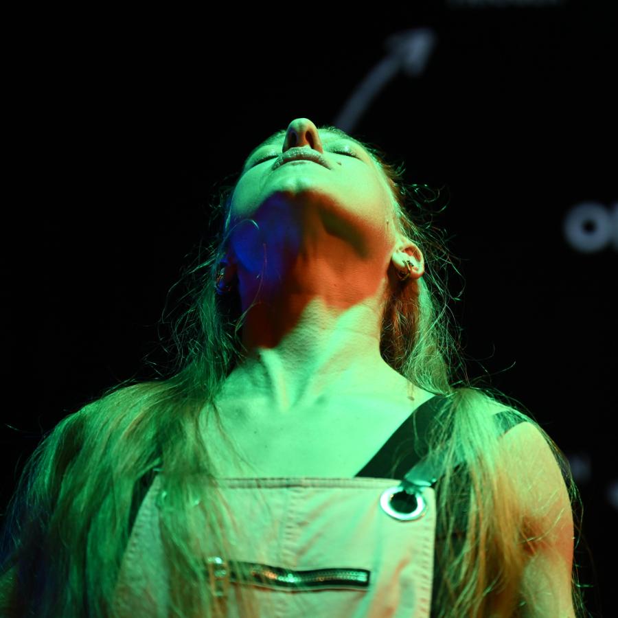 Image credit: Skyler Reid. A woman with long hair stands on stage with her head thrown back, highlighted by an intense green light. 