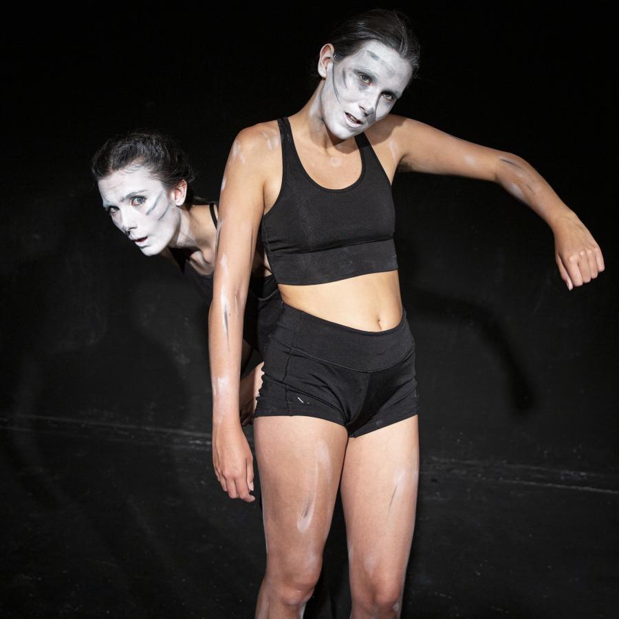 Two woman, their faces smeared with white paint and black lines, one peeking behind the other. 