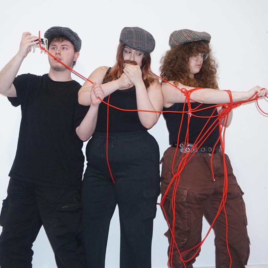  Three people wearing flatcaps all tangled in red rope. One person is looking at a plastic toy cow, another is lighting a cigarette, and another is looking at the rope tangled on their arm. 