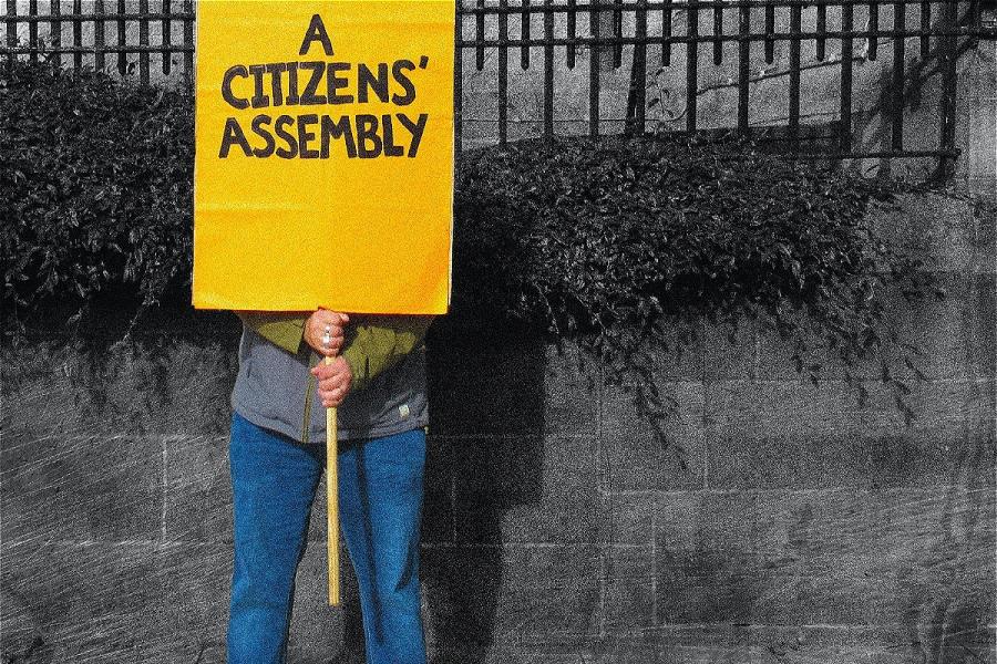 A person holds an orange placard that says A CITIZENS' ASSEMBLY