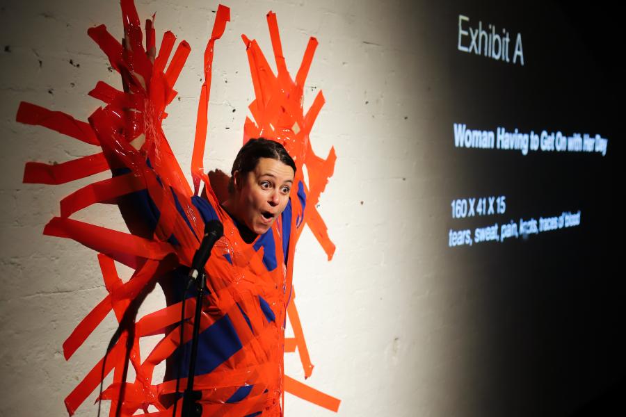 A woman wearing a bright blue jumper is taped to a white wall with bright red tapes. She is speaking into a microphone with an expression between surprise, pain and amusement. We distinguish some words projected on the back wall aking to a museum artwork 