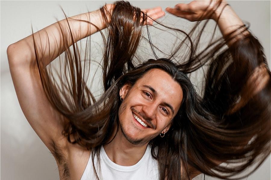 Alexis, the performer, wears a white tank top and poses with their arms above their head, their hair draped in all directions, in front of a white wall, their head tilted and a bright smile on their face.