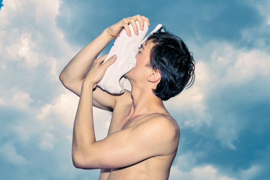 A shirtless young man sniffs a white trainer against the background of a blue sky, the sun shining bright behind him.