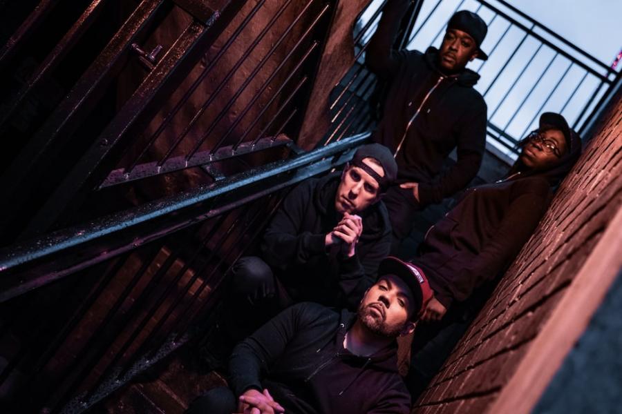 Four people in hoodies and caps sit and stand on a metal staircase inbetween 2 brick walls. They all have straight sturn expressions looking at the camera. The view is angled to the side.