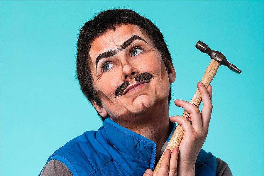 Shoulders up portrait of drag king: 'Dad'. Dad wears a grey t-shirt and a bright blue gilet, he is holding a thin hammer lovingly in his hands. Dad looks dreamily off camera, he has short dark hair and painted on facial hair. Light blue background.