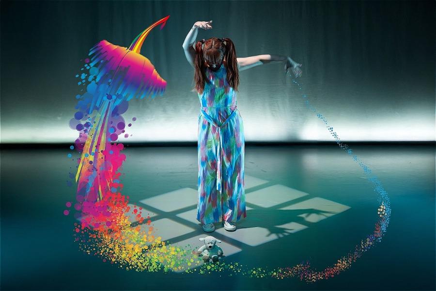 A woman in a colourful dress stands on a stage, her arms extended, casting a shadow. A stuffed toy lies on the floor in front of her. Vibrant rainbow effects form a phoenix shape around her.