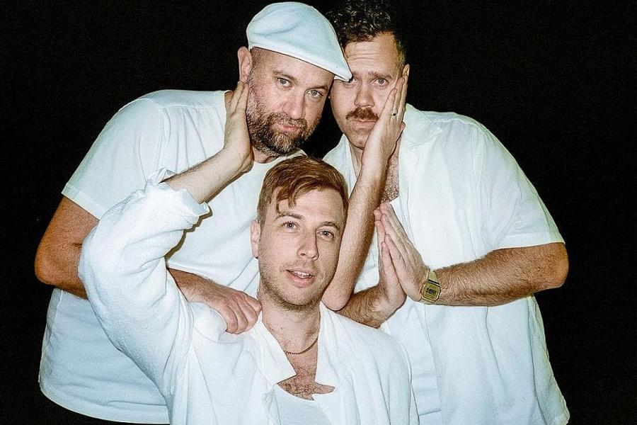 Three caucasian men in white against a black background, positioned in an inverted triangle. They are variously touching each other's cheeks and shoulders.