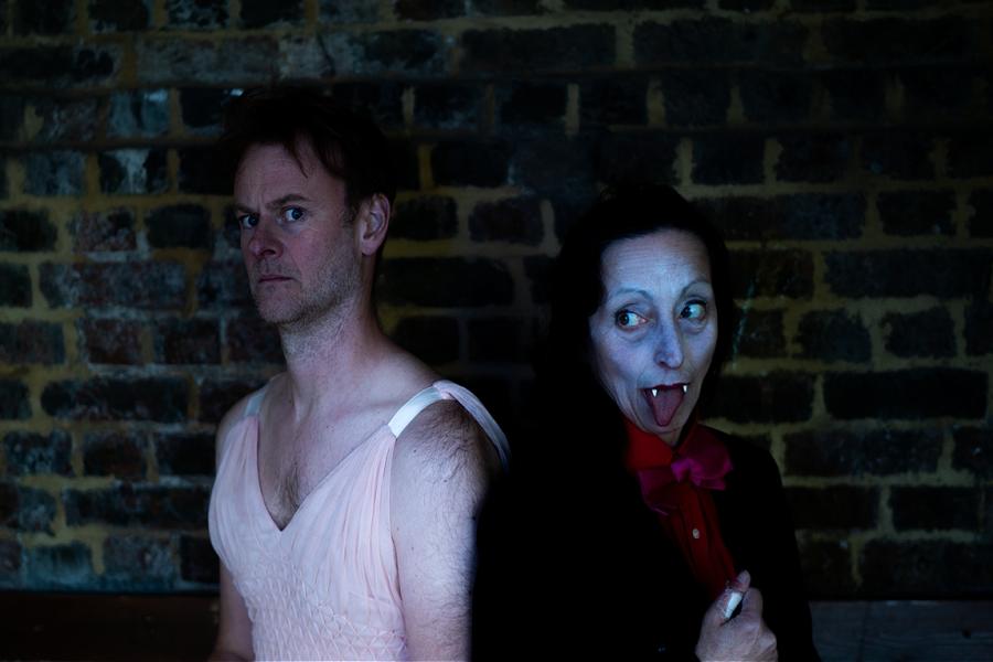 A woman with fangs, dressed in a black dinner jacket and red shirt. A man wearing a pink dress. They are sat back to back looking at the camera. Behind them is a brick wall.