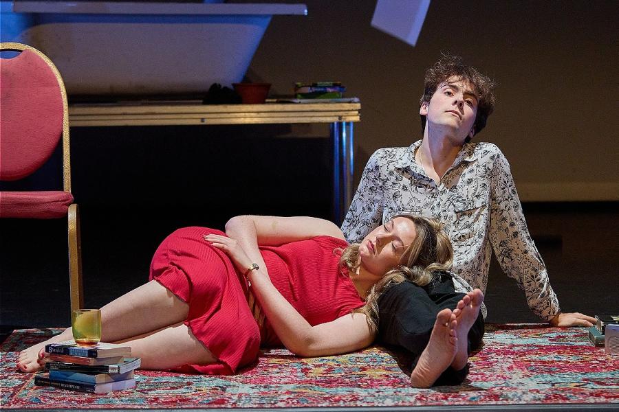 Two Actors. One in a white shirt and black trousers is sat with their legs infront of them, one laid down across their lap wearing a red dress. There are books next to them, a bathtub behind them and sheets of paper falling onto the floor.