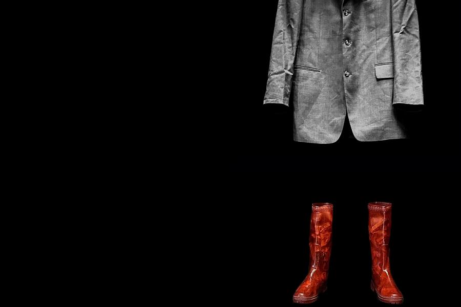 A grey suit hangs above a pair of red wellies on a black background.