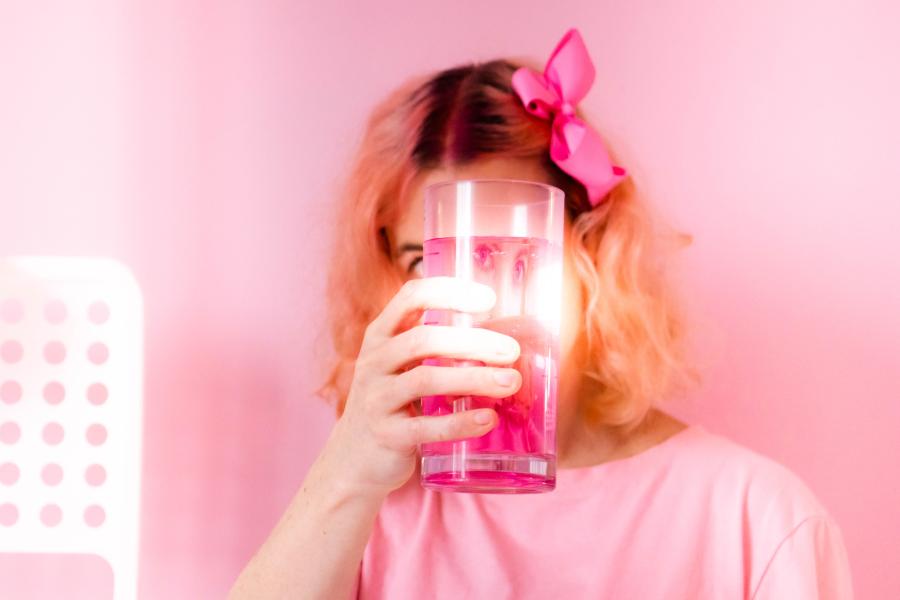 Head and shoulders of a white woman dressed all in pink against a pink wall. She is holding up a glass with pink liquid in front of her face so that it is only visible distorted, through the reflection in the glass's domed sides.