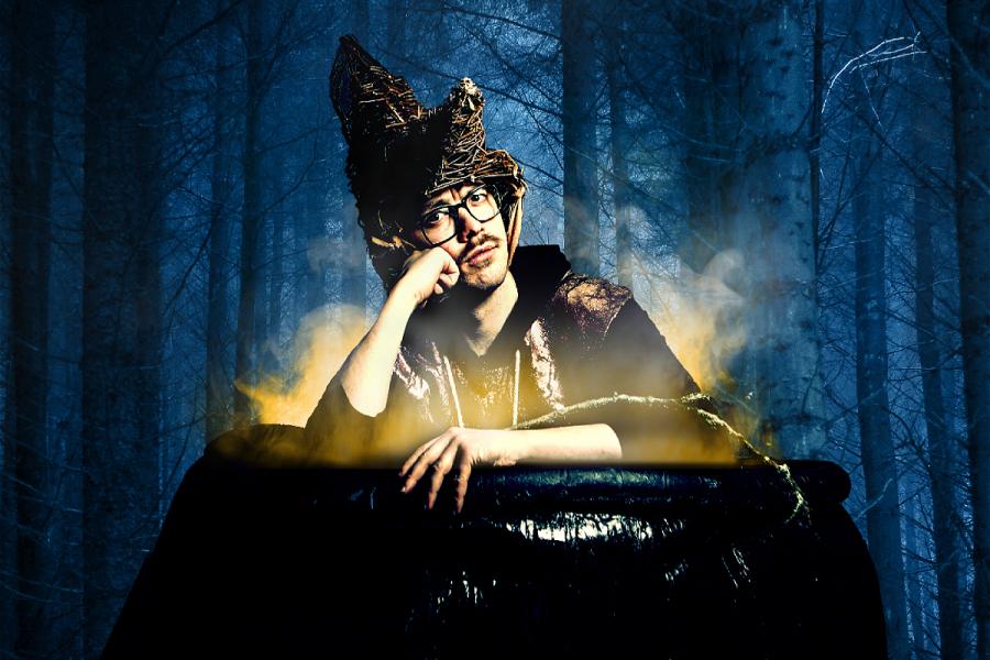 A man in a robe with black thick rimmed glasses and a moustache looks fed up in a giant DIY cauldron with a folk-ish spooky wicker hat on his head. In the background is a wood of trees with a dark blue background.