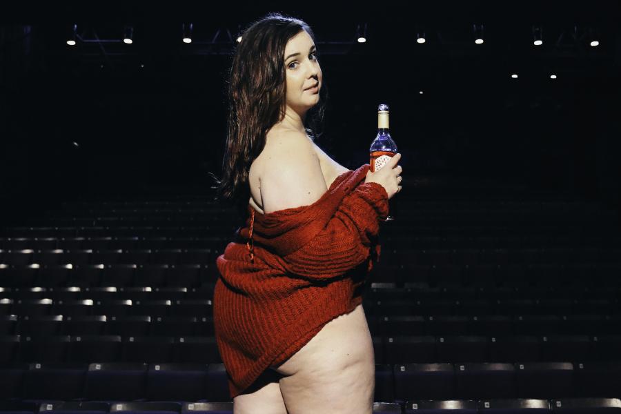 A fat woman semi-naked with a red cardigan draped over her shoulders, looks back into the camera with a bottle of wine in her hand. She stands on a stage.