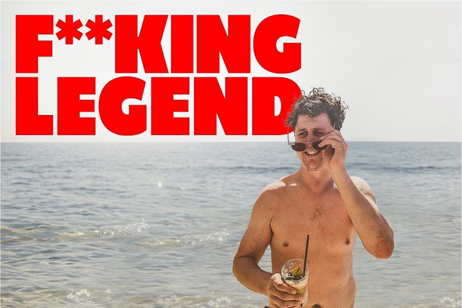 The protagonist stands on a sunny beach, removing his sunglasses, holding a drink that's almost spilling, he looks confused. In large red capital letters, the words F**KING LEGEND appears above him.