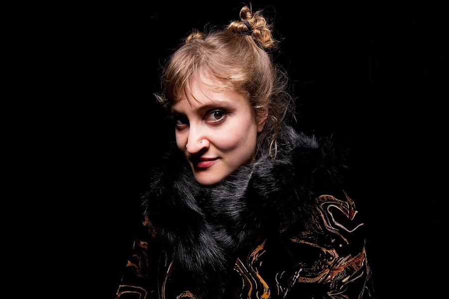 Frankie is a blonde, dark eyed white femme presenting person against a black background. She has two buns in her hair looking suggestively down the camera as if to say 'I dare you'. She is wearing some interesting faux fur fabrics that drown her.