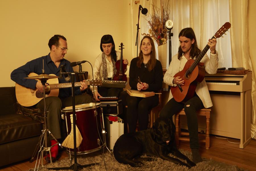 Four people, from left to right - Tom Adams, Claire Orme, Siobhan Meade and Dan Scott sitting in a lounge playing musical instruments such as guitar, xylophone and with a piano behind. Marty, the guide dog, is laying on the floor and both Siobhan and Mart