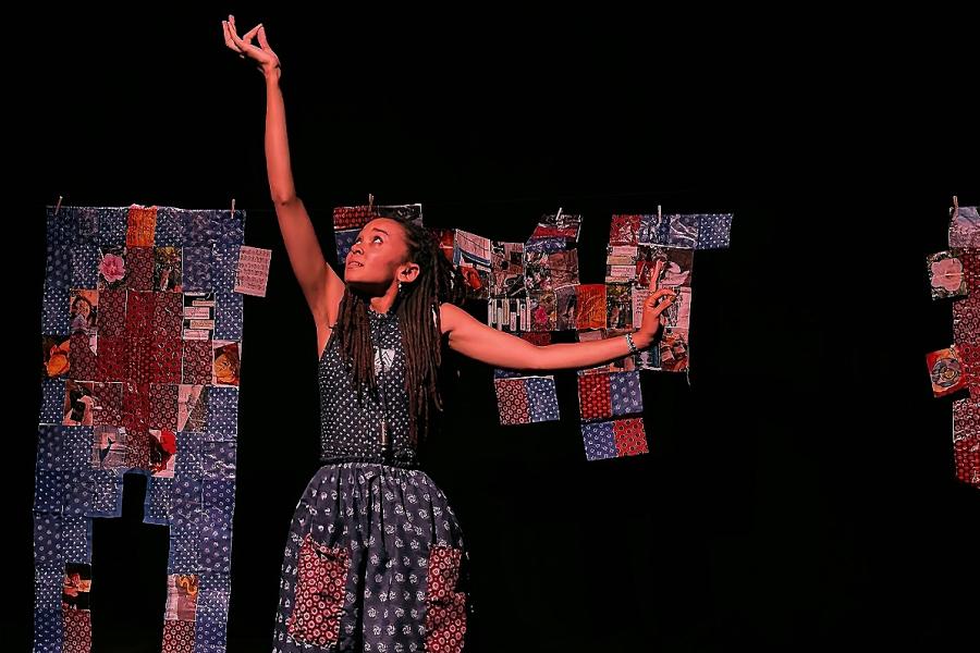 A performer stands on a dark stage with one arm raised and the other extended, gazing upward. She wears a blue patterned dress with red patchwork pockets, echoing the quilt panels hanging behind her on clotheslines.