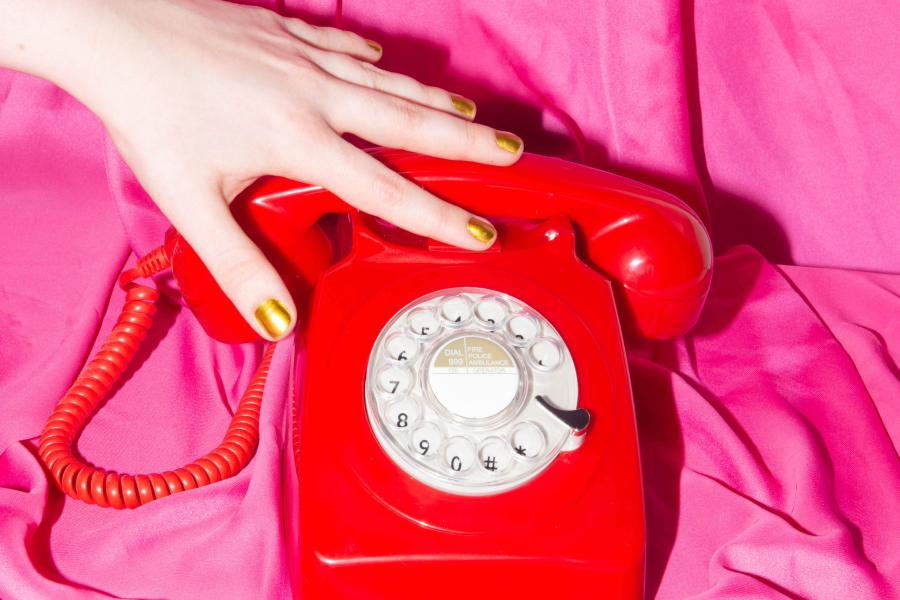 A red 80s style telephone is placed against a pink silky material backdrop. A white woman's hand with gold nail vanish, has her hand placed on top of the phone, as if she is stroking it.
