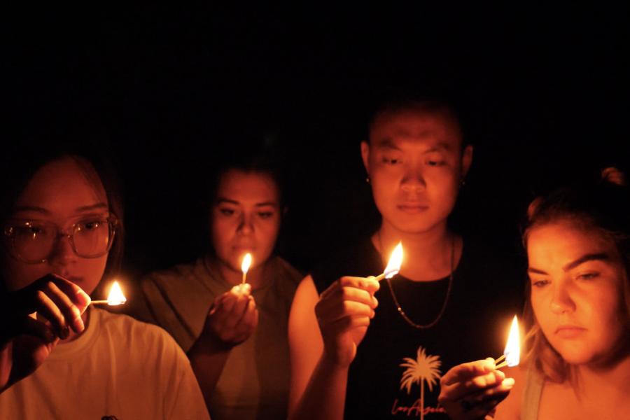 Four people stand facing the camera a lit match in their hand. It is dim. The only light in the image is from the light from the small flames. From left to right; Yuanfan Pan is an Asian woman with brown hair and brown eyes, Roshi Cowen is a medium build 