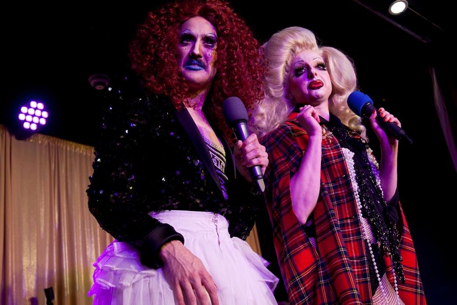 Two drag people stare ahead disparagingly.  The left hand person wears a red wig and black sequinned jacket. They glare through blue lipstick.  The right hand person purses their red lips.  They both hold microphones and stand in front of a gold curtain.