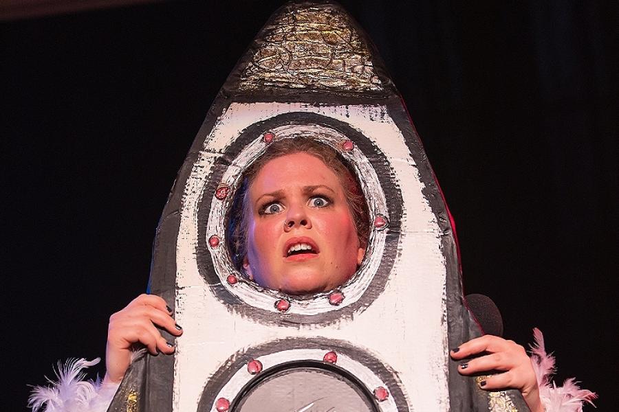 A performer dressed with their head poking out of a cardboard cut out of a rocket ship.