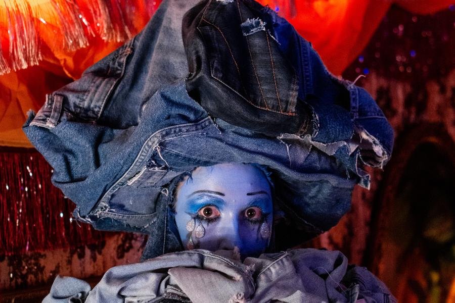 Drag thing, Jean, pokes their face out of a pile of denim