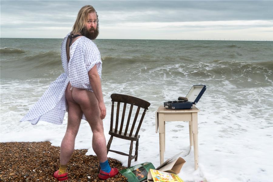 James (white blond, bearded man) stands in the wash of waves from the sea wearing a medical gown with his bum out. next to him is an empty chair and a table with a record player on it. On the ground are discarded records, the sea and sky are grey