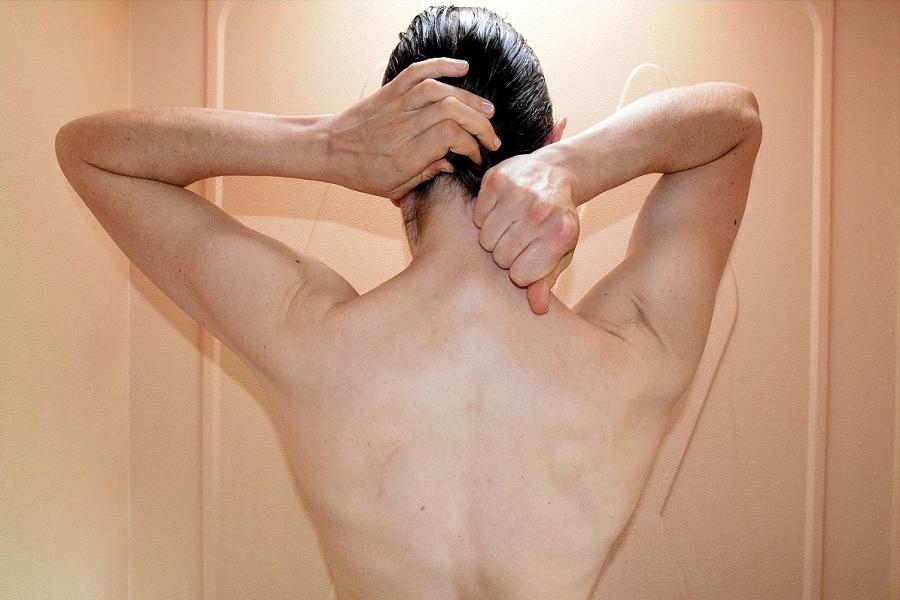 Close up of the strong naked back of a white person in a shower. They are scraping their dark brown hair back into a low ponytail.  Their gender is unidentifiable.  Their arms and fists are strong.