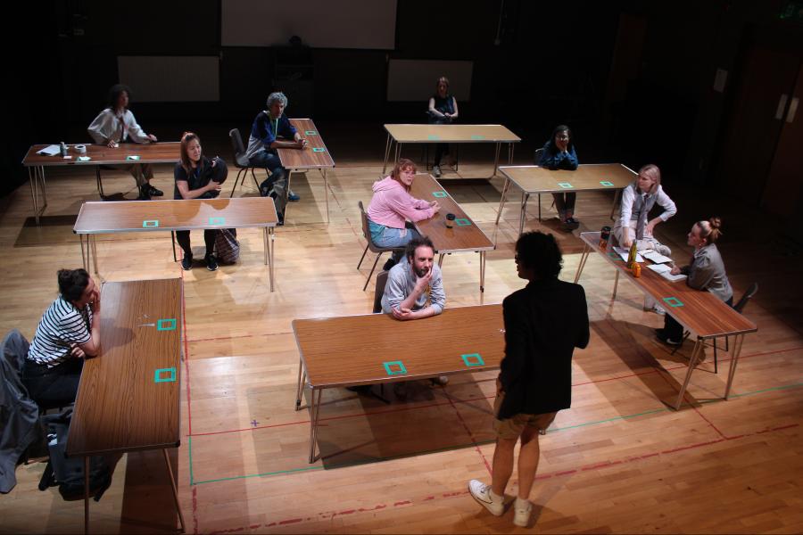 Landscape - view from above, a group of desks arranged in right angles. Audience members are sitting at the desks as one of the performers stands at the front explaining a task, holding a stack of post-its in his hands.  Portrait - two performers frantica