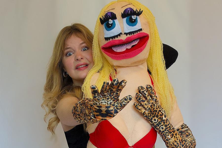 A woman sits with a life-sized puppet woman on her knee. Both are blonde and wearing long leopard print gloves. The puppet wears a red bra and smiles at the camera. The woman behind her wears all black and looks panicked, reaching towards the viewer.