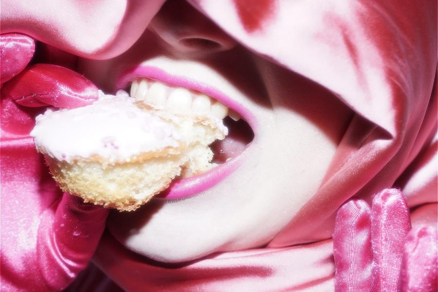 LINDA is shrouded in folds of pink fabric. They wear dark pink gloves and have bright pink lipstick on. They are biting into a small pink cupcake. Their teeth are on show tearing off a piece of sponge.