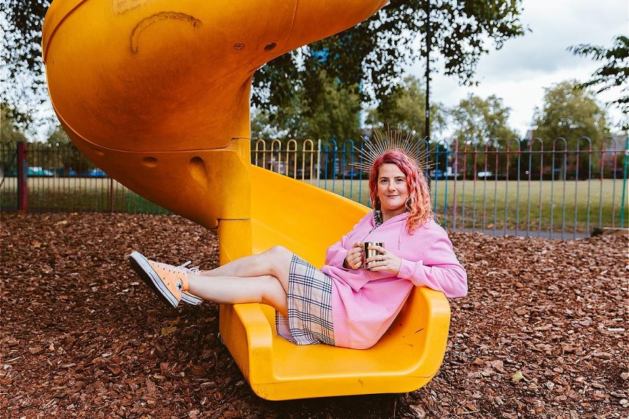 Kelly Green is a 40 year old white woman who has bright pink and orange hair. She is wearing a checked print dress that is similar to the Burberry print, a bubblegum pink hoody, and neon orange Converse.. They are wearing a halo headband that is made of g