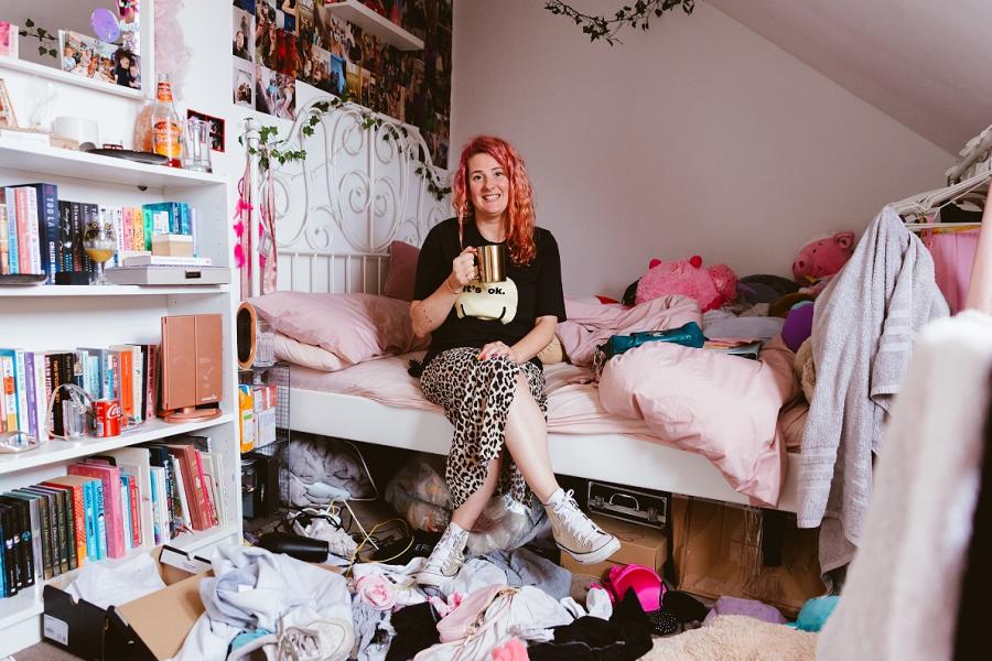 Kelly is sitting on a bed with a gold mug in a messy teenage girls bedroom. Kelly has pink hair & is smiling uncomfortably. She wearing a black t-shirt with a yellow smiley face, its eyes spelling out 