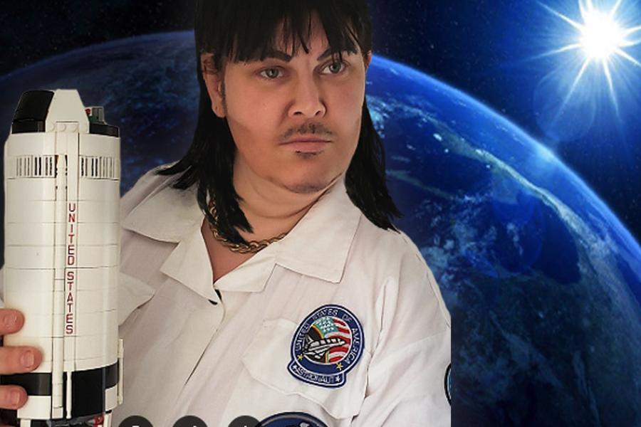 A drag king dressed in a white boiler suit with a black mullet. They are holding a Lego model of the Apollo 11 rocket. There is a starfield in the background. The text says Max Velocity Takes Space.