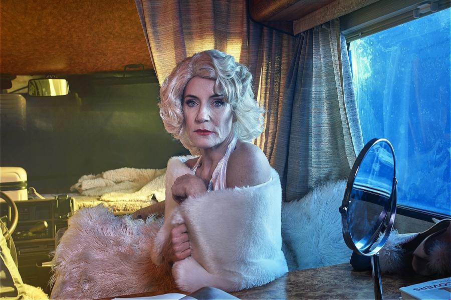 An older woman dressed as Marilyn Monroe poses inside a dimly lit motorhome. She wears a platinum blonde wig, a white halter dress reminiscent of Monroe's iconic look, and a white fur stole. She sits at a wooden table with an open photo book in front of h