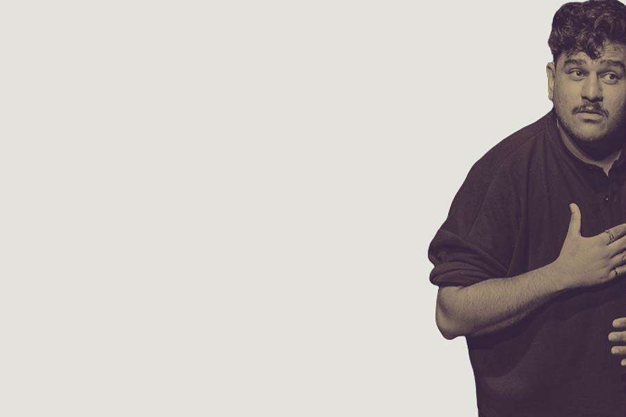 A Brown Deaf Man, stocky build wearing a black top, sleeves up. With black hair fluffed to one side, black moustache on its own, eyes wide open as he look like he's asking a question to the audience with both hands on the chest as a BSL for 'patience'