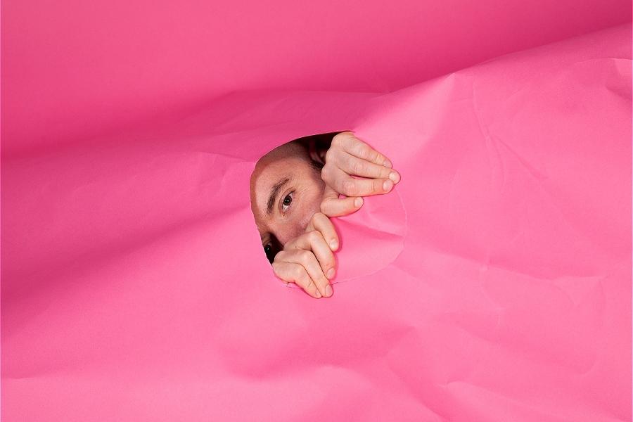 A person peeking through a tear in a large pink sheet of paper.