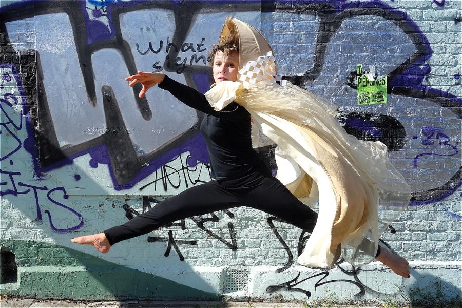 A woman in a cloak jumps into the air, in front of a graffitied wall.