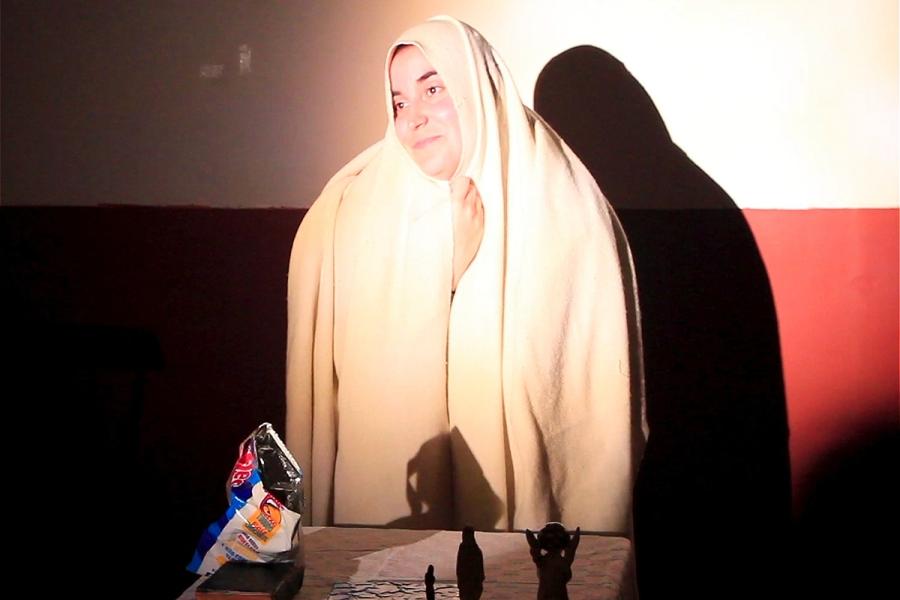 A woman, wearing a white veil, alluding to the Virgin Mary, smiling, standing behind a table. On top of the table, a bible and an opened bag of crisps.