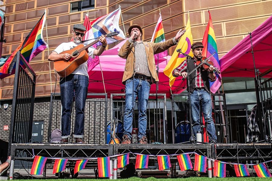 Three men in their 40s and early 50s, playing a guitar, accordion and violin, are performing in front of pride flags. The accordion player, who is wearing a bwoler hat, is also singing. The other two men are in caps.
