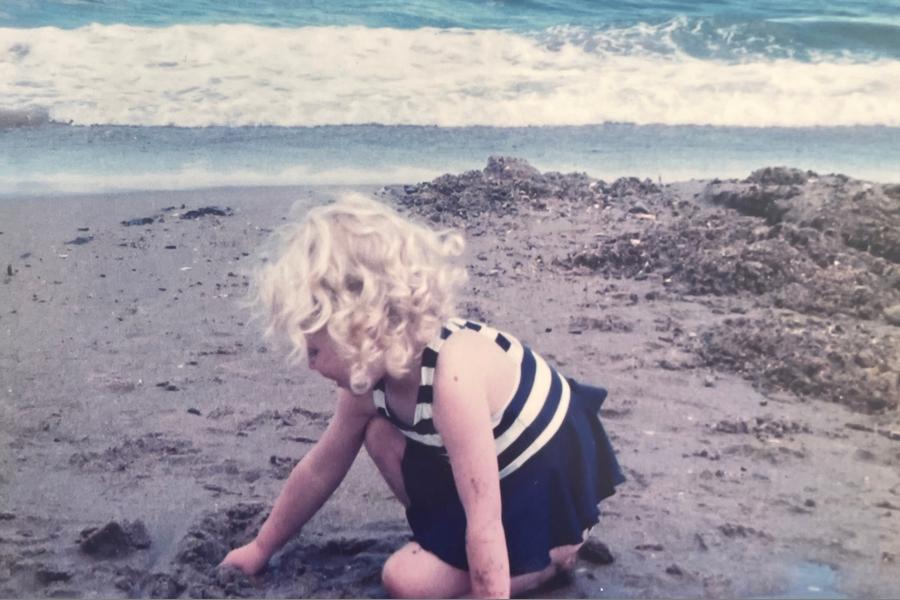 A nostalgic old photograph: the colour is slightly faded. In it, a young girl with curly blonde ringlets, wearing a black and white swimming costume, is on the beach. She is below the horizon, playing with the sand in her chubby hands. 